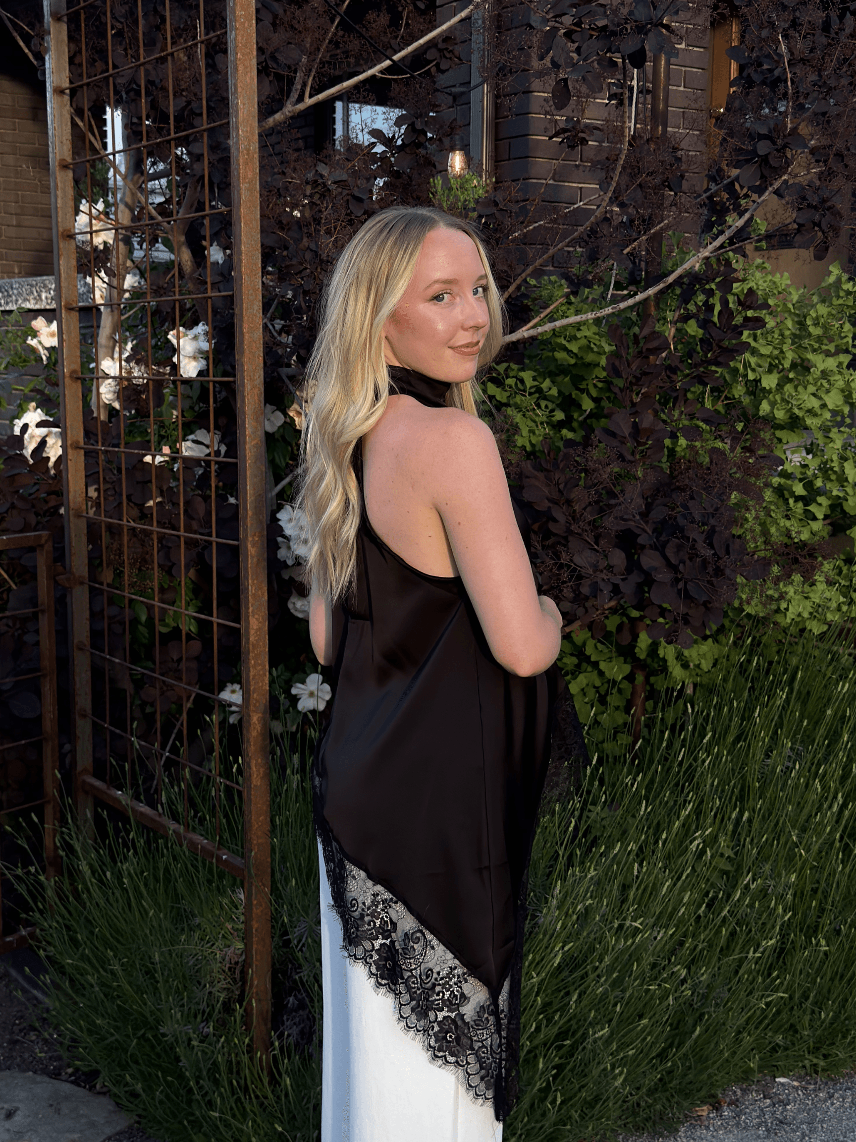 Blonde woman in a black satin top with lace trim poses in a garden.