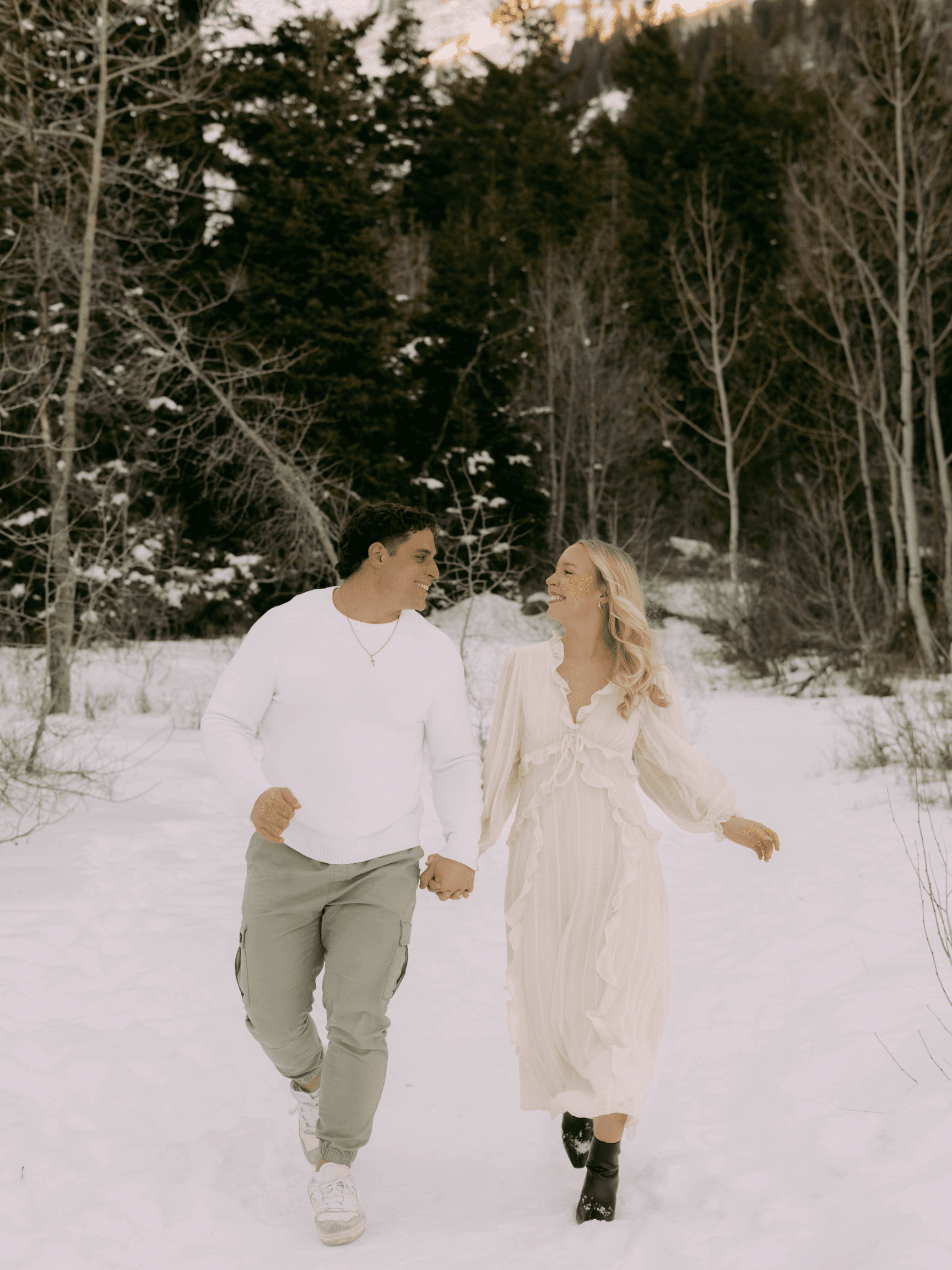 Smiling couple walks hand-in-hand through a snowy forest, wearing white and light green winter attire.
