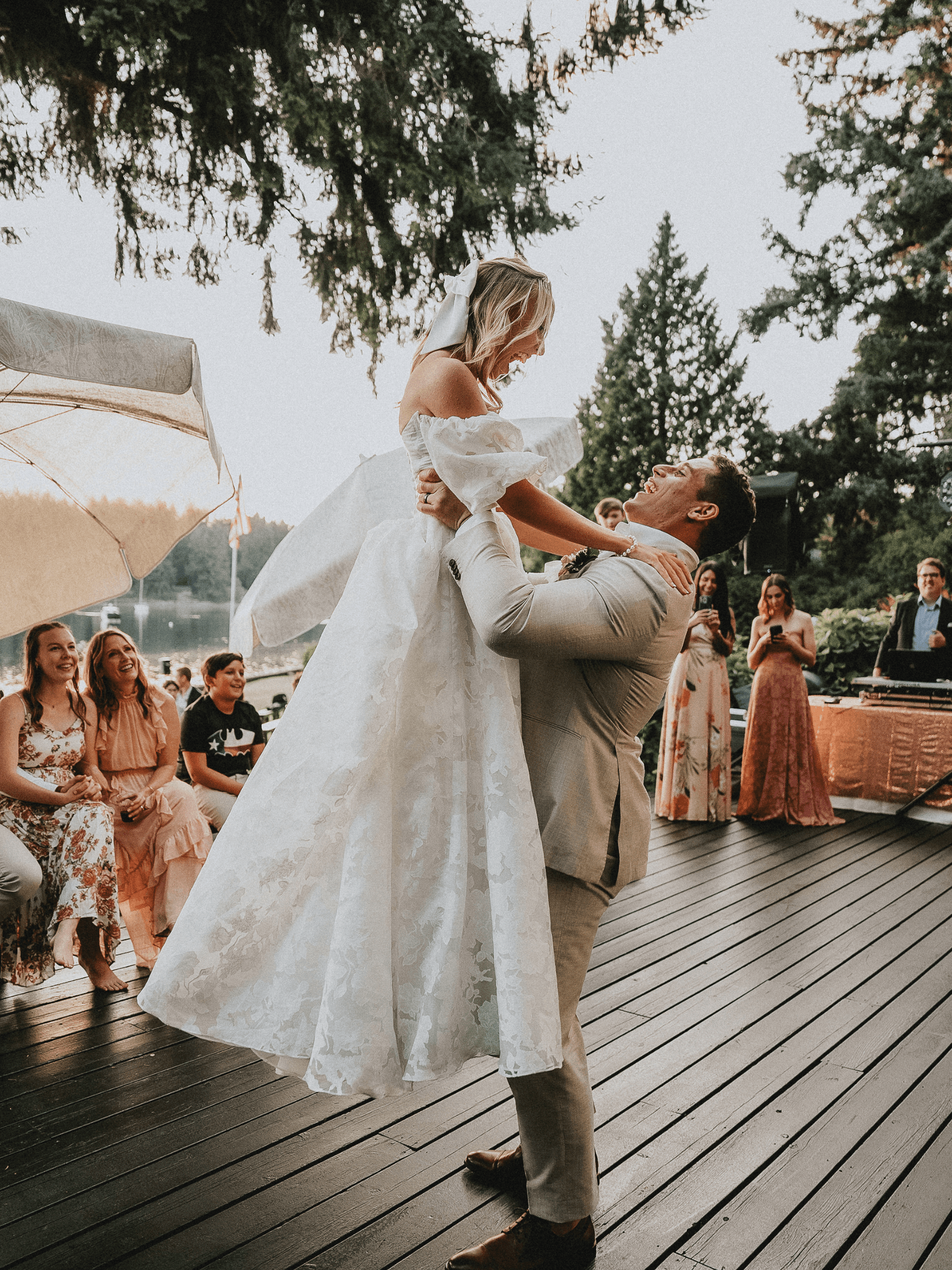 Groom lifts bride in white gown during outdoor wedding reception on a wooden deck.